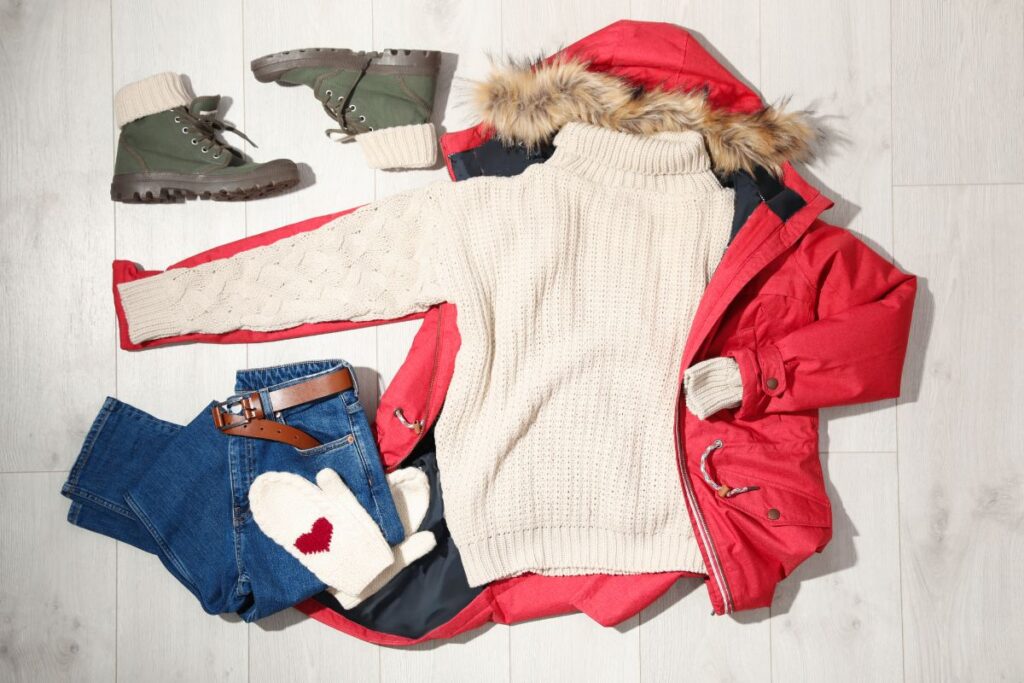“Flat lay of winter clothing including a red parka, cream sweater, jeans, boots, and knit mittens.”